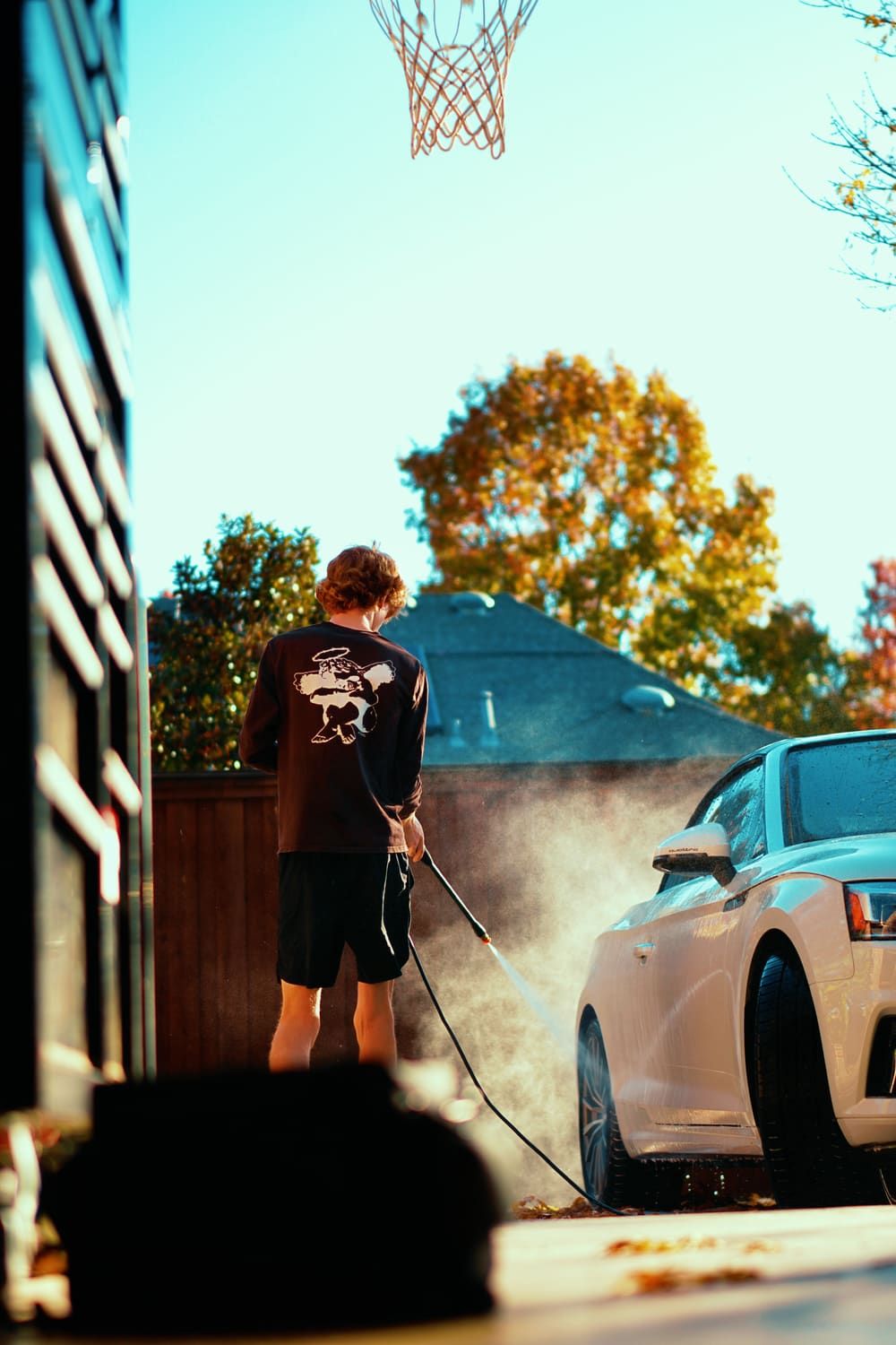 A person seen from behind pressure washing a light-colored car in a driveway, with a basketball hoop and autumn trees behind.