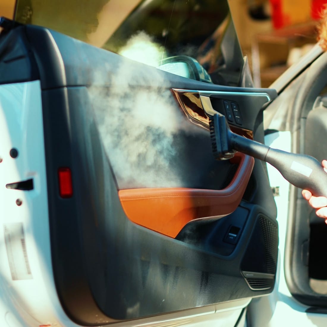 Steam rising off a car's interior door panel as it is steam cleaned, next to tan leather trim.